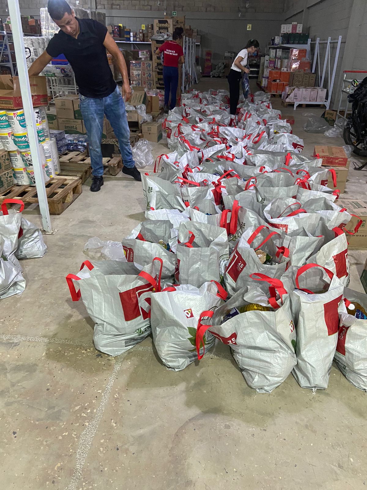 Warehouse scene with a man arranging boxes and volunteers packing rows of grocery bags with red handles