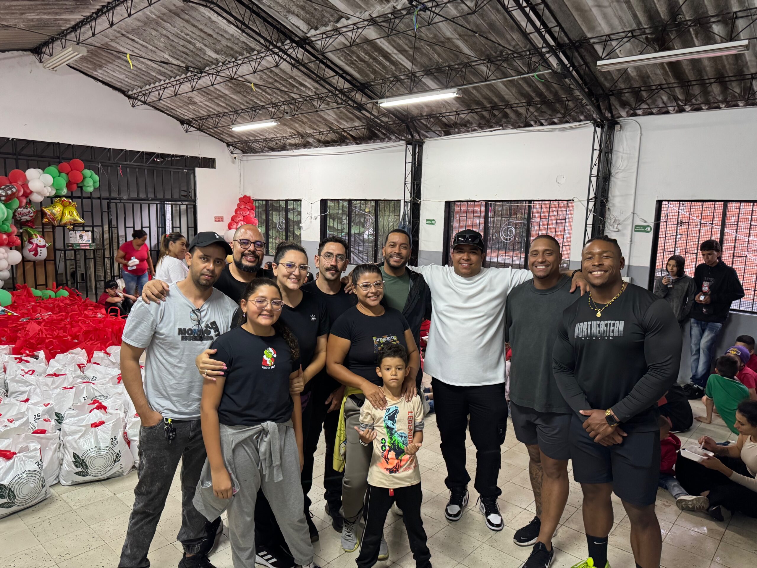 Large group of volunteers and children posing indoors beside stacks of gift bags and Christmas balloons